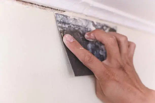 Photo of A handyman applies putty or filler to cover the gap between a wood cornice and a concrete wall with a trowel. Home improvement or renovation.