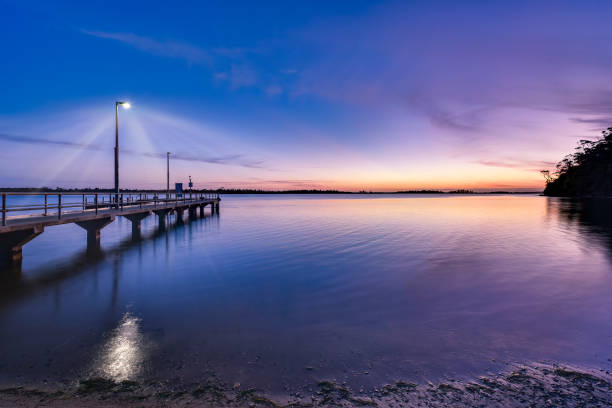 Walkway with a railing, fence lines at pier, entrance leading to the sunset in the evening time over lake, silhouette across water and reflection of clouds on calm water.