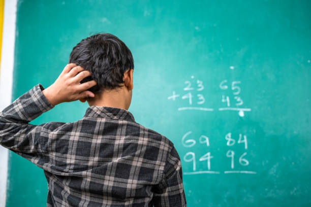 confused kid scratching head to solve mathematics problem on board at classroom - concept of intelligence, trouble learning and education - matemática imagens e fotografias de stock
