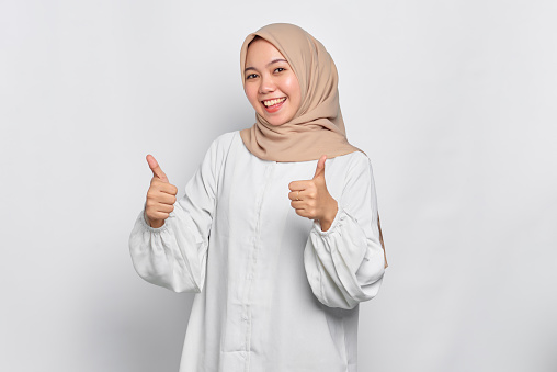 Smiling young Asian Muslim woman showing thumbs up gesture isolated over white background