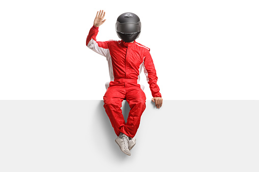 Full length portrait of a racer with a helmet sitting on a blank panel and waving isolated on white background