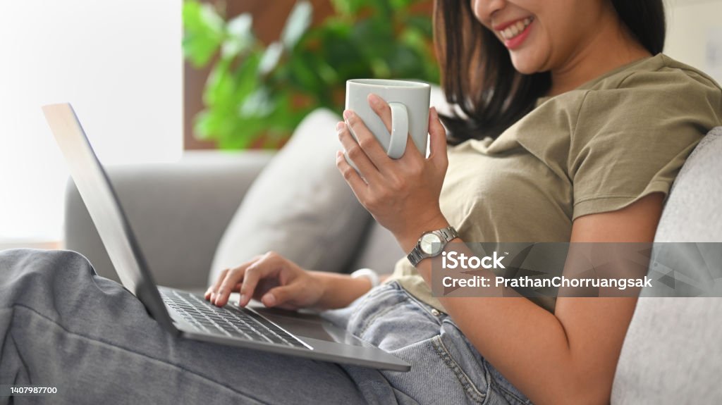 Cheerful young woman holding cup of coffee and surfing internet on laptop computer. 20-29 Years Stock Photo