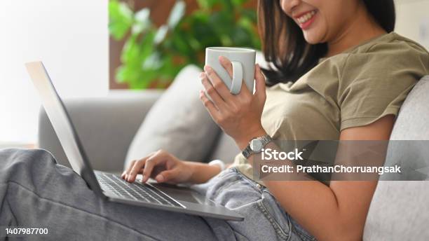 Cheerful Young Woman Holding Cup Of Coffee And Surfing Internet On Laptop Computer Stock Photo - Download Image Now