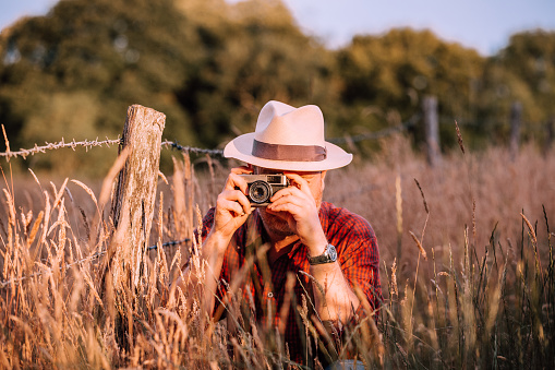 Portrait of a male photographer in his 30s shooting with a retro film camera that uses 35mm film. He is wearing a panama hat, red check shirt and is surrounded by rural nature and golden tall grass on a summer evening.