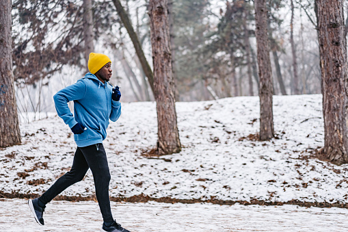 Young handsome black man jogging outdoors in forest during winter, on the snow.