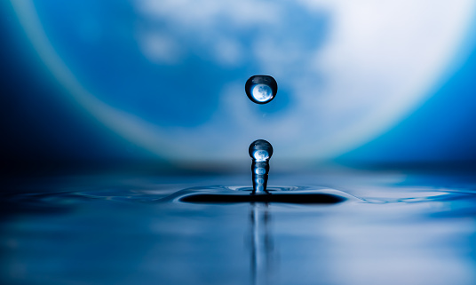 A close-up of a water drop on the surface of the water with a full moon in the background and reflecting on the water and refracting in the drop.