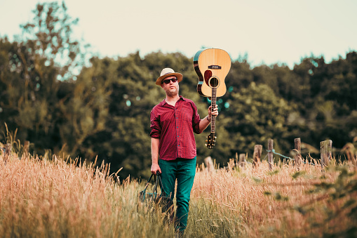 A man walks alone on a rural path surrounded by golden summer meadows and tall grass. He carries an acoustic guitar, leather bag and he wears a red check shirt, panama hat and sunglasses. Room for copy space.