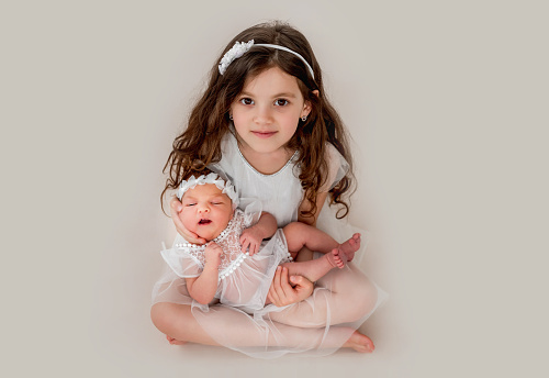 Beautiful little girl sitting, looking at the camera and holding her beautiful sleeping newborn baby sister. Studio portrait of adorable napping kid with her sibling together