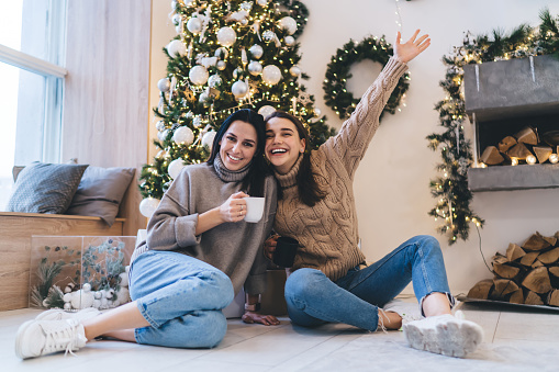 Full body of cheerful young women in comfy outfits cuddling and drinking hot   coffee while celebrating New Year together at home sitting against decorated Christmas tree