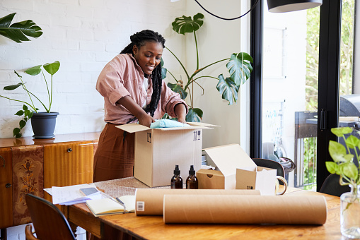 Smiling young African woman standing at a table and packing orders from her online home-based business into boxes for shipping