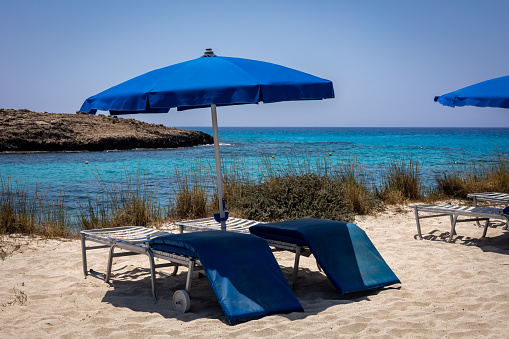 An umbrella and two empty sunbeds with blue mattresses on the sandy beach with a sea view. Nissi beach, Cyprus.