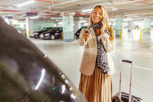 Cropped shot of a young businesswoman walking on the underground parking and carrying suitcase. Young woman with luggage opening the trunk in parking garage, talking on the smartphone.