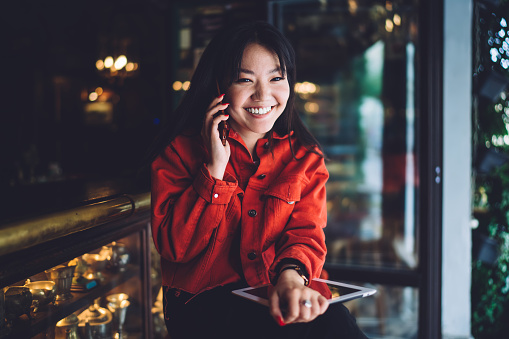 Happy young ethnic female with dark hair in red jacket smiling while having phone conversation sitting in modern outdoor cafe with tablet