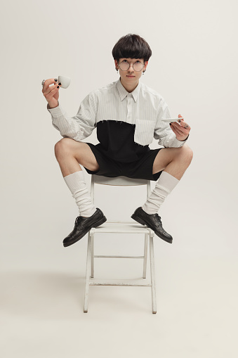 Portrait of stylish young man with piercing sitting on chair, drinking coffee isolated over grey studio background. Concept of modern fashion, art photography, style, queer, uniqueness, ad