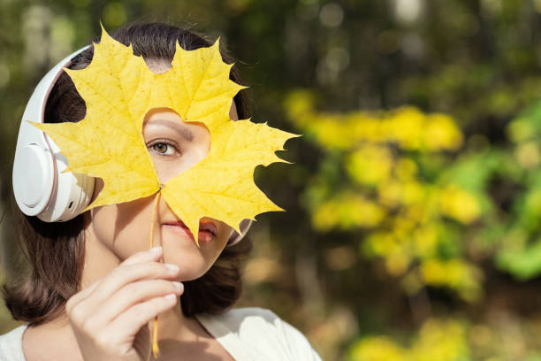 piękna szczęśliwa młoda kobieta w słuchawkach zamykająca twarz z żółtego liścia klonu i patrząca przez dziurę w kształcie serca w jesiennym parku - maple leaf audio zdjęcia i obrazy z banku zdjęć