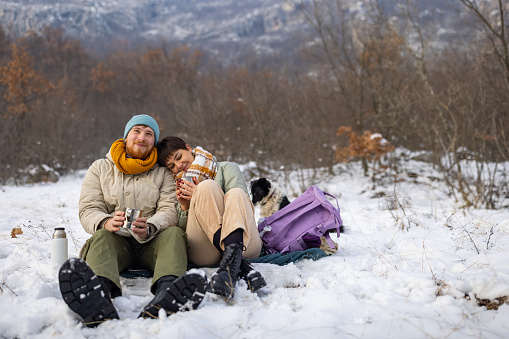 Young Caucasian couple, enjoy the winter day adventure at the mountain, while relaxing after hiking, and drinking tea while dog makes them a company