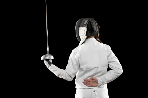 Defender moving. One sportsman, female fencer in white fencing costume in basic stance isolated on black background. Sport, youth, hobbies, achievements, goal. Girl practicing with rapier
