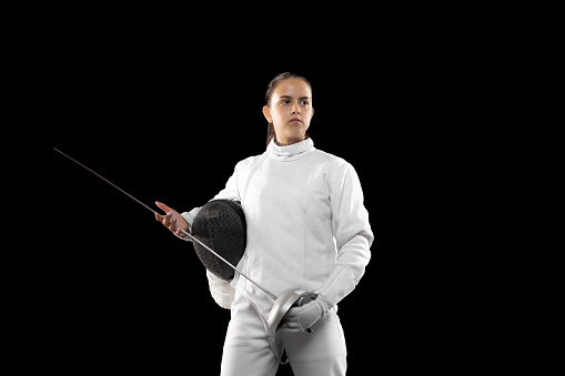Young serious girl, beginner fencer in white fencing costume standing with rapier ans mask isolated on dark background. Sport, youth, activity, skills, achievements. Solo