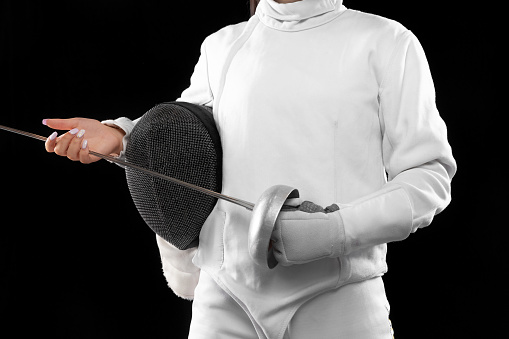 Closeup female fencer's body in white fencing costume with mask and rapier isolated on dark background. Sport, youth, activity, skills, achievements.