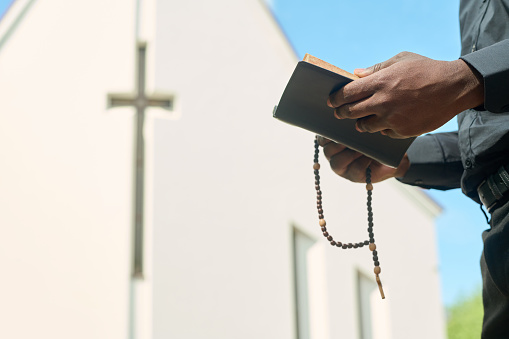 Hands of priest in black apparel holding rosary beads and open Gospel while reading verses and explaining them during sermon