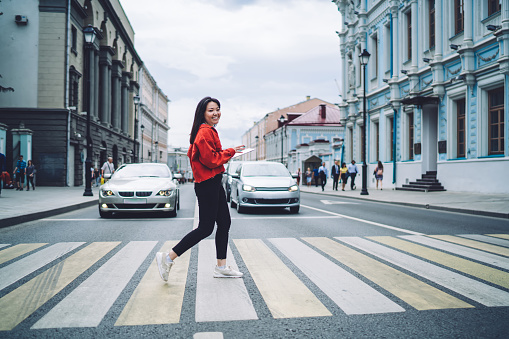 Side view of positive young Asian female pedestrian in red jacket using tablet while crossing road against modern cars in city district