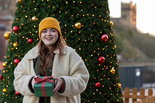 A young female adult standing in front of a Christmas tree receiving a wrapped present with a red bow and smiling looking into the camera. There is a cathedral out of focus in the background.