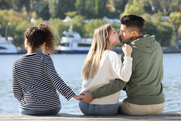 mężczyzna trzymający się za ręce z inną kobietą za plecami swojej dziewczyny na molo w pobliżu rzeki. trójkąt miłosny - passion kissing black white zdjęcia i obrazy z banku zdjęć