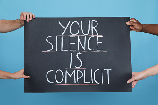 People holding sign with phrase Your Silence Is Complicit on light blue background, closeup. Racism concept