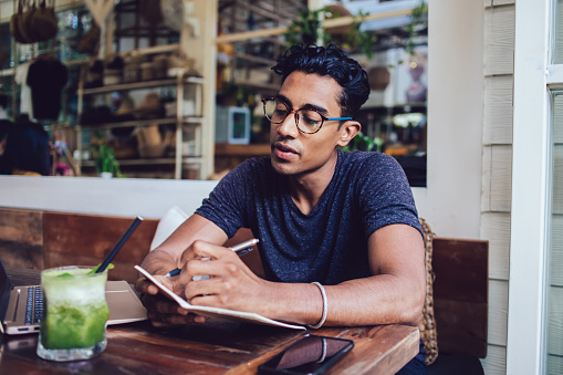 Wistful focused Hispanic male chilling at wooden table in cafe while making notes in copybook and relishing cooling beverage on blurred background