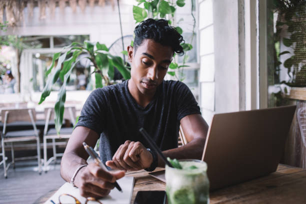 Serious ethnic man looking at wrist watch while working and using laptop at terrace Thoughtful ethnic man in casual outfit checking time browsing laptop and sitting at table with notebook and glass of exotic drink at street cafe time management student stock pictures, royalty-free photos & images