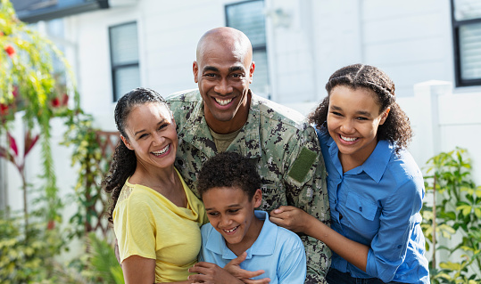 Portrait of a proud African-American navy veteran with his multiracial family, standing together outside their home. The two children and their mother are African-American and Caucasian. The parents are in their 40s, their daughter is 12 years old and their son is 8. All except the boy are smiling at the camera.