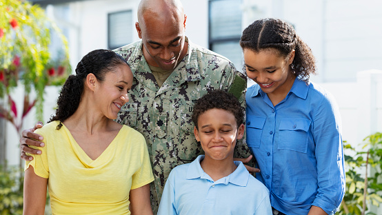 An African-American navy veteran with his multiracial family, standing together outside their home. The two children and their mother are African-American and Caucasian. The 8 year old boy is making a face and everyone is looking down at him. The parents are in their 40s, and their daughter is 12 years old.