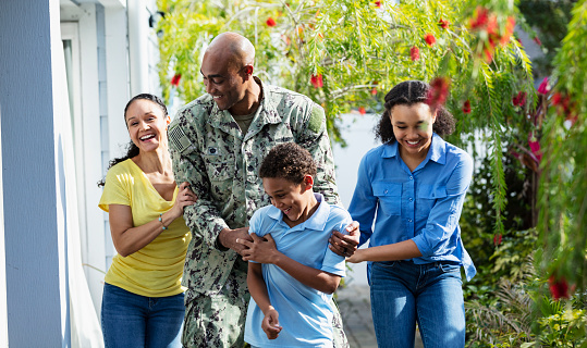 An African-American navy veteran with his multiracial family, walking together outside their home on a sunny day, smiling and laughing. The two children and their mother are African-American and Caucasian. The parents are in their 40s, their daughter is 12 years old and their son is 8.