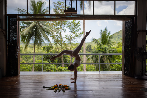 Active and flexible young woman in silhouette, doing a yoga handstand pose, at the yoga center, with view of rainforest