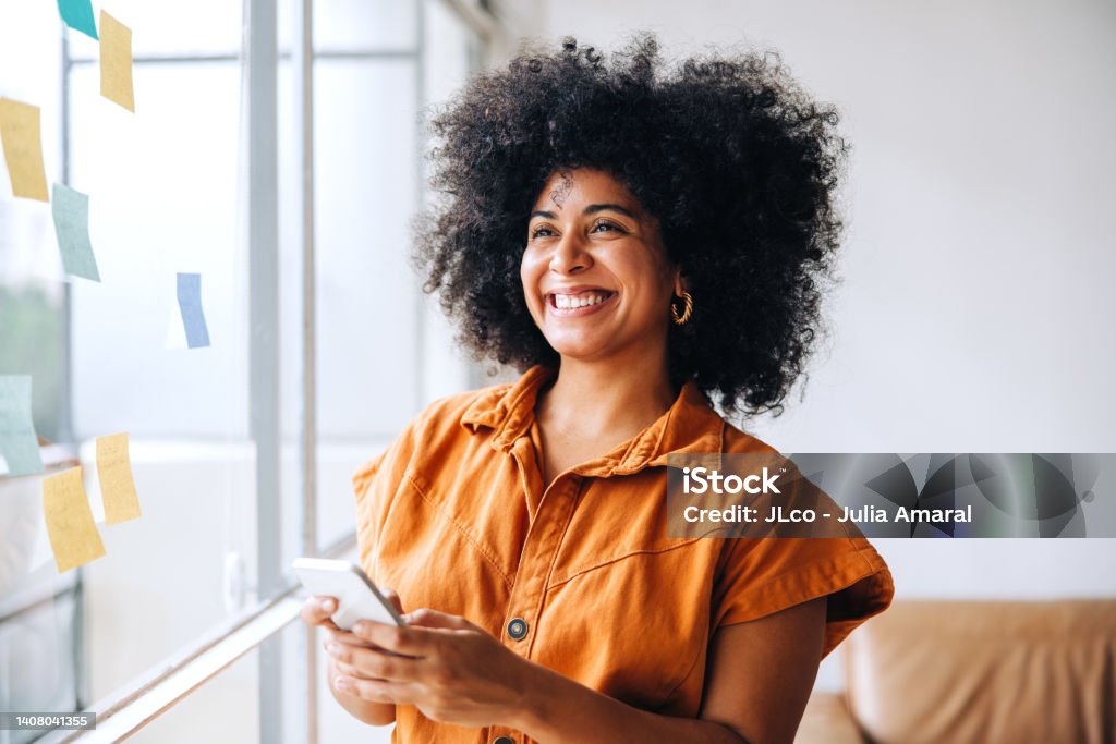 Happy black businesswoman using a smartphone in a creative office Happy black businesswoman smiling while using a smartphone in a creative office. Cheerful female entrepreneur sending a text message while standing next to a glass wall with sticky notes. Women Stock Photo