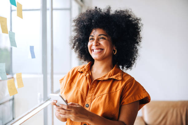 Happy black businesswoman using a smartphone in a creative office Happy black businesswoman smiling while using a smartphone in a creative office. Cheerful female entrepreneur sending a text message while standing next to a glass wall with sticky notes. millennial generation stock pictures, royalty-free photos & images