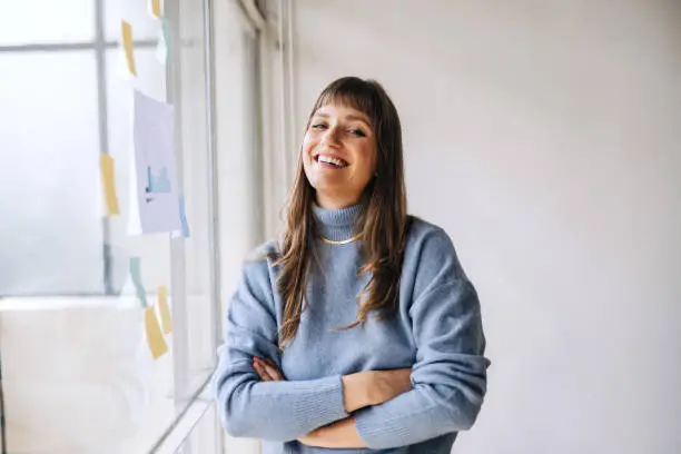 Cheerful young businesswoman smiling at the camera while standing next to a glass wall in an office. Happy female entrepreneur organising her creative ideas using adhesive notes.