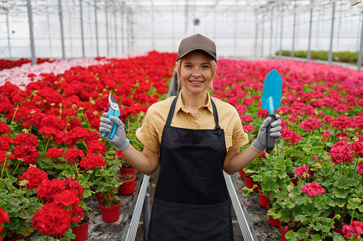 Portrait of a cheerful female greenhouse worker with a gardening tool in hands. Female florist with pruner and trowel