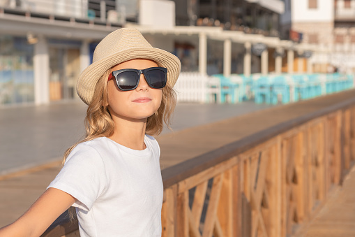 A cute young girl in a hat and sunglasses stands on a wooden promenade.