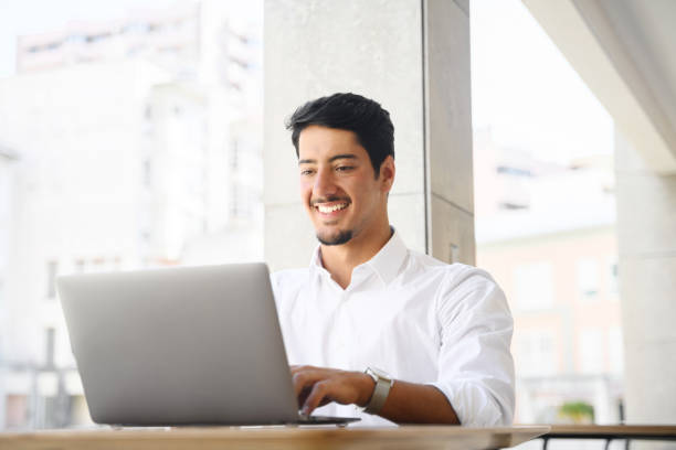 Latin male developer, freelancer, office employee is using laptop sitting at the desk oudoors. Young multiracial guy is typing on the keyboard stock photo