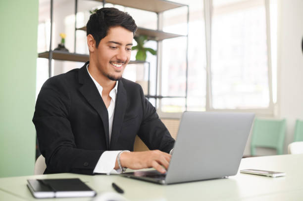 istock Optimistic ethnic man in formal business wear using laptop sitting on the workplace in modern office 1408049649