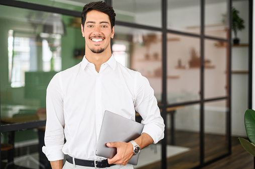 Working with joy. Smiling cheerful hispanic business man wearing formal white shirt standing with laptop