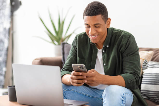 istock Successful man entrepreneur or freelancer sitting on sofa in home office chatting using smartphone. Young student male working, using mobile phone and laptop, smiling 1408049897