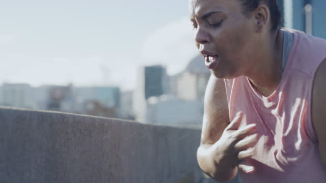 A plus size woman with chest pain while exercising in the city. Exhausted and unfit female suffering from asthma, breathing heavily and hitting her chest after a cardio workout along a highway
