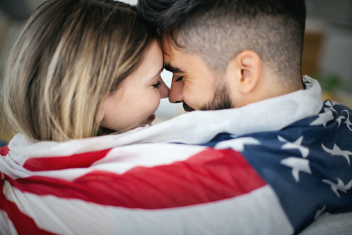 Young couple in love wrapped in American flag, looking at each other, about to kiss.