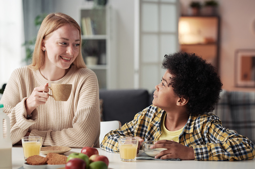 Young happy mother drinking coffee and talking to her adoptive son at table during breakfast