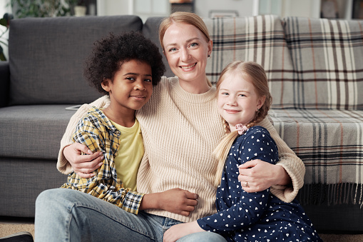 Portrait of young happy mother embracing her adopted children and smiling at camera sitting on floor in the room