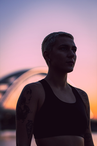 Portrait of a young tattooed woman with short hair on an early morning outside workout. Determined and disciplined young woman out for a run, during a beautiful sunrise by the river and a bridge.