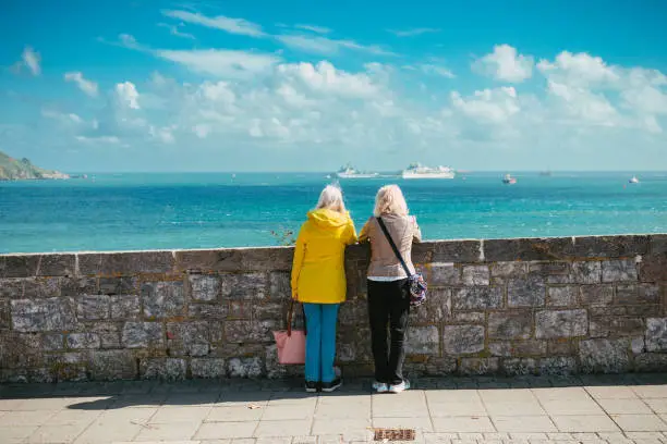 Color image depicting the rear view of two senior women in their 70s looking at the sea and harbour on a sunny summer day. Blue sky and cloudscape, with boats defocused in the distance.