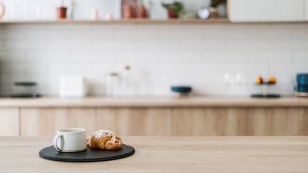 Croissant and coffee on kitchen countertop, against blurred interior Croissant and coffee on kitchen countertop, against blurred minimalist interior with modern furniture. Selective focus at homemade pastry and tea drink in cup on wooden table, copy space, web banner kitchen counter stock pictures, royalty-free photos & images